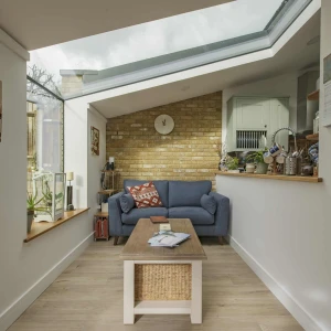 A cozy living room featuring a blue sofa, a wooden coffee table, and a brick wall. Large windows allow natural light to fill the space, and there are various plants and decorative items on display. A clock and shelves can be seen in the background.
