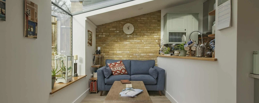 A cozy living room featuring a blue sofa, a wooden coffee table, and a brick wall. Large windows allow natural light to fill the space, and there are various plants and decorative items on display. A clock and shelves can be seen in the background.