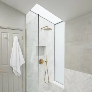 A modern bathroom featuring a glass shower enclosure with a gold showerhead and fixtures. The walls are tiled with light-colored marble in a herringbone pattern. A white towel hangs on the door. Natural light enters through a skylight above the shower.