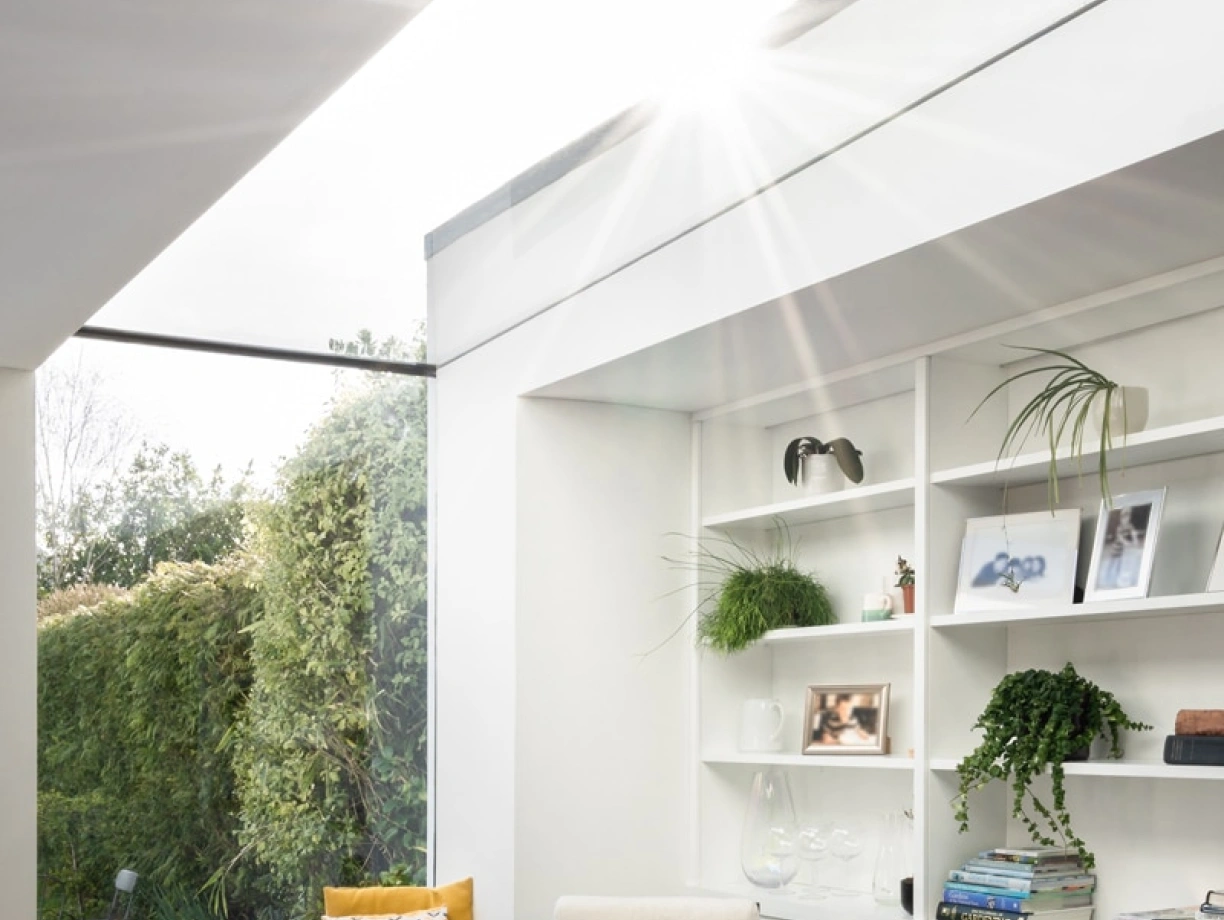 Bright, modern living space with a cozy armchair and a stylish bookshelf. Sunlight streams through a skylight, illuminating greens from outdoor plants visible through a glass wall. A comfortable nook features a patterned cushion against a window.