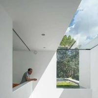 A man leans on a white balcony railing, looking out towards a glass window that frames a view of trees and the sky. The interior has clean lines and minimalistic design. Natural light creates striking shadows on the walls.