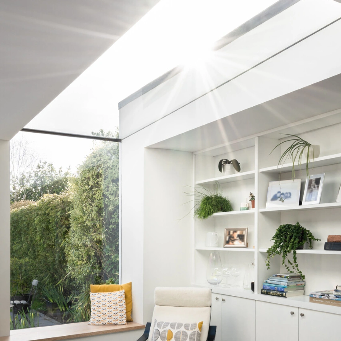 Bright, modern interior featuring a cozy chair by a window, with built-in white shelving adorned with plants and decorative items. Sunlight streams through a skylight, illuminating the space. A lush garden is visible outside.