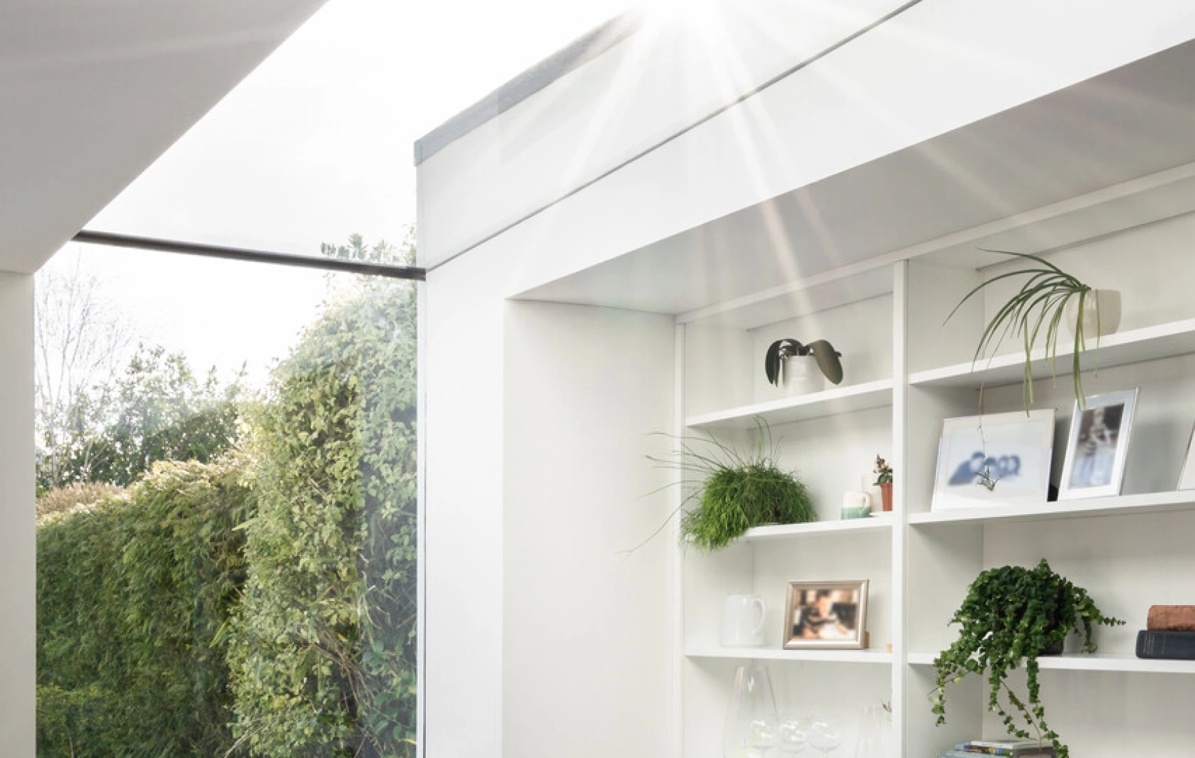 Espacio luminoso con un sillón blanco y negro, estanterías con decoraciones y plantas. Ventana amplia que permite la entrada de luz solar y vistas al jardín exterior. Suelo de madera clara.