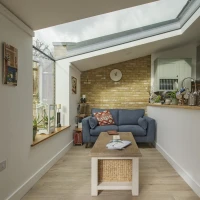 Un salon lumineux avec un canapé bleu, une table en bois clair et un mur en briques apparentes. Des plantes sont disposées sur des étagères, et un grand velux laisse entrer la lumière naturelle. Un décor moderne et accueillant.