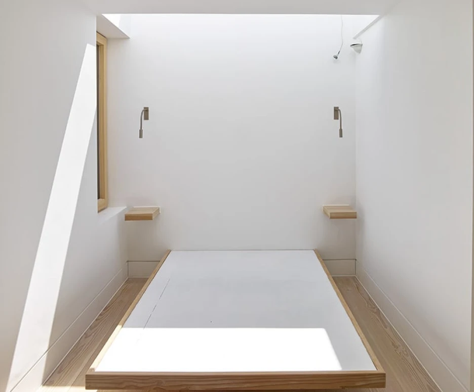 A minimalist bedroom with a wooden platform bed centered in the room. Natural light streams in through a large window, highlighting the white walls and light wooden flooring. Two wall-mounted lamps are positioned on either side of the bed.