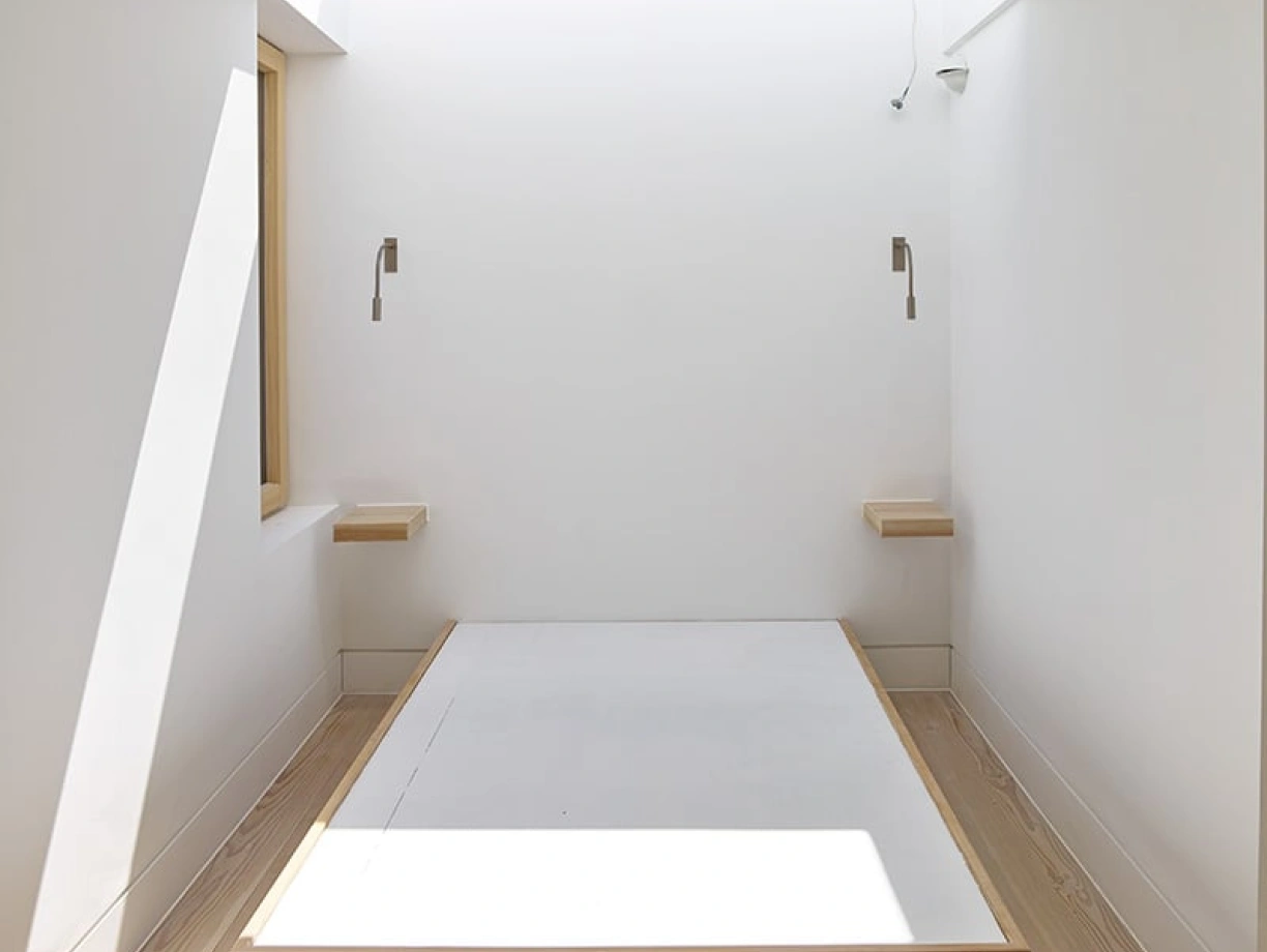 A minimalist bedroom with a wooden platform bed centered in the room. Natural light streams in through a large window, highlighting the white walls and light wooden flooring. Two wall-mounted lamps are positioned on either side of the bed.
