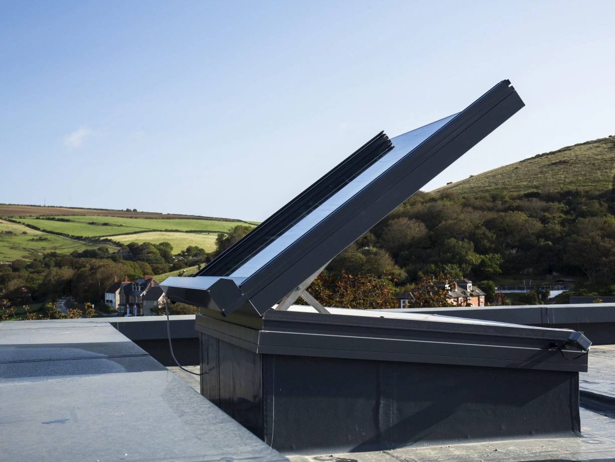 A solar panel array mounted on a sloped rooftop, angled to capture sunlight. The background features lush green hills and clear blue skies.