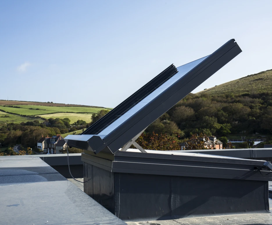 A solar panel is installed on a rooftop, angled to capture sunlight, with a scenic view of hills and greenery in the background. The sky is clear and blue.