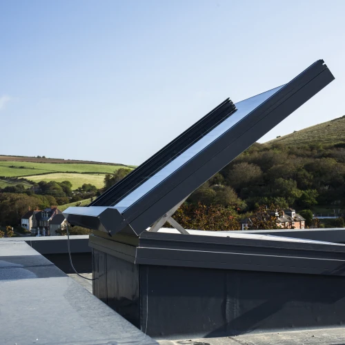 A solar panel is installed on a rooftop, angled to capture sunlight, with a scenic view of hills and greenery in the background. The sky is clear and blue.