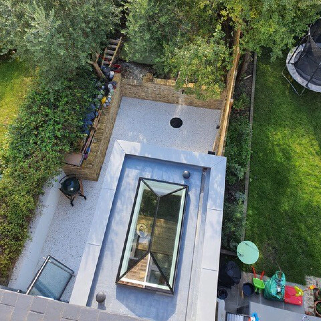 Vista aérea de un patio con un área de césped, un camino de piedra y un estanque circular. Se observa un tragaluz en el techo de la casa. Al fondo, árboles y un trampolín.
