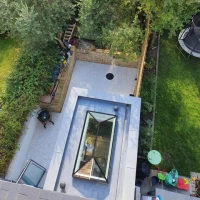 Vista aérea de un patio con un área de césped, un camino de piedra y un estanque circular. Se observa un tragaluz en el techo de la casa. Al fondo, árboles y un trampolín.