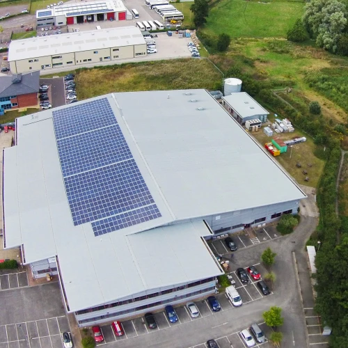 Vue aérienne d’un bâtiment industriel avec des panneaux solaires sur le toit, entouré de verdure et d’un terrain de stationnement. Des installations supplémentaires sont visibles à proximité, ainsi que des champs.