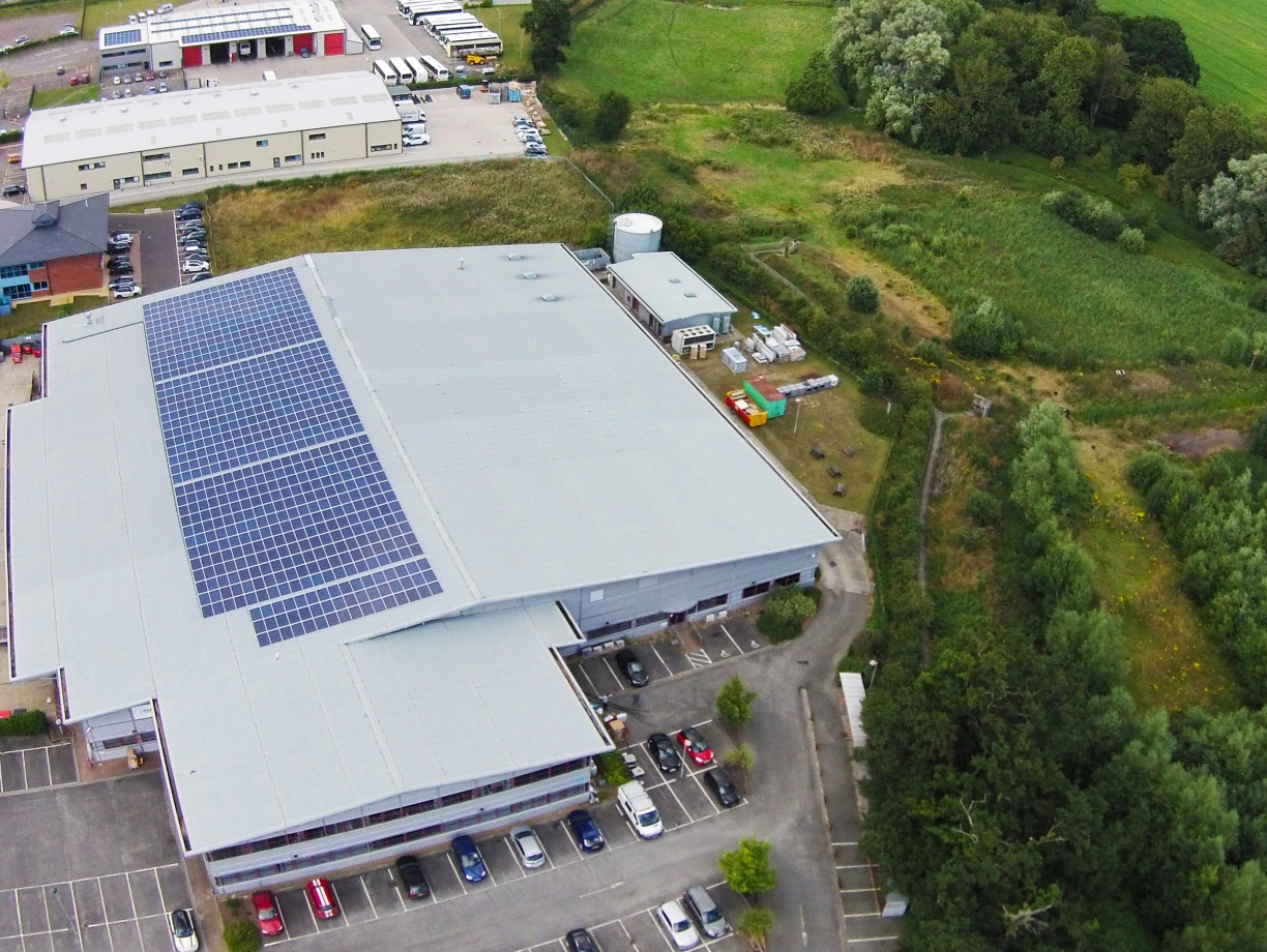 Aerial view of a large commercial building with a rooftop solar panel array. The building is surrounded by a parking lot filled with cars, green fields, and trees in the background. A small structure and some equipment are visible beside the building, blending into the natural landscape.