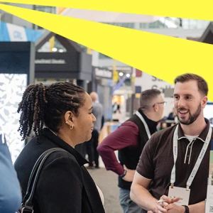 A diverse group of people engage in conversation at a trade show or exhibition. A man is speaking animatedly to a woman while others listen intently. Colorful banners and booths are visible in the background.
