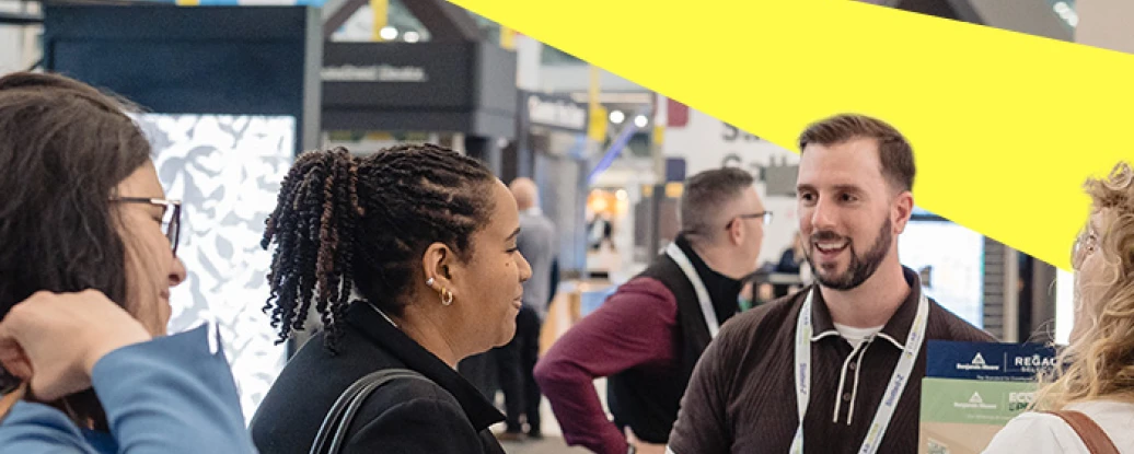 A diverse group of people engage in conversation at a trade show or exhibition. A man is speaking animatedly to a woman while others listen intently. Colorful banners and booths are visible in the background.