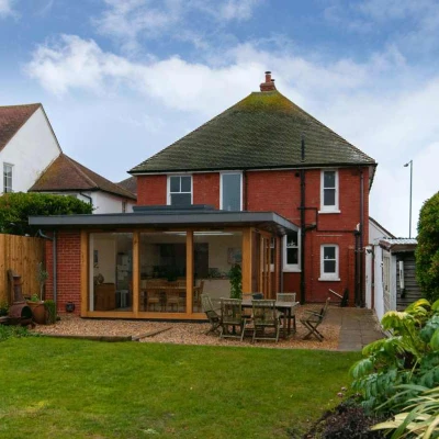 A red brick house with a modern glass extension in the backyard, surrounded by greenery and a wooden fence. The lawn is neatly manicured, and there is outdoor furniture on a paved area.