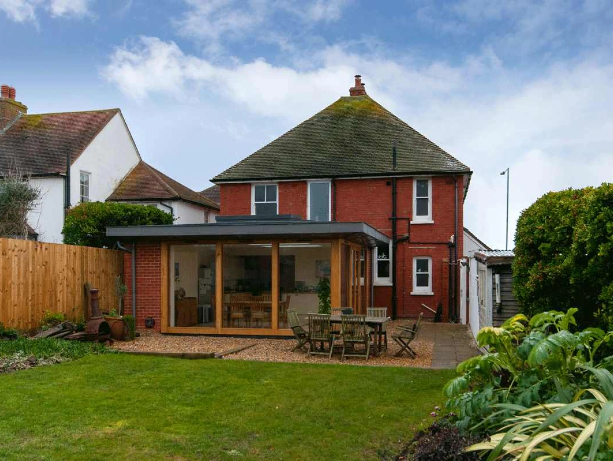 A red brick house with a modern glass extension in the backyard, surrounded by greenery and a wooden fence. The lawn is neatly manicured, and there is outdoor furniture on a paved area.