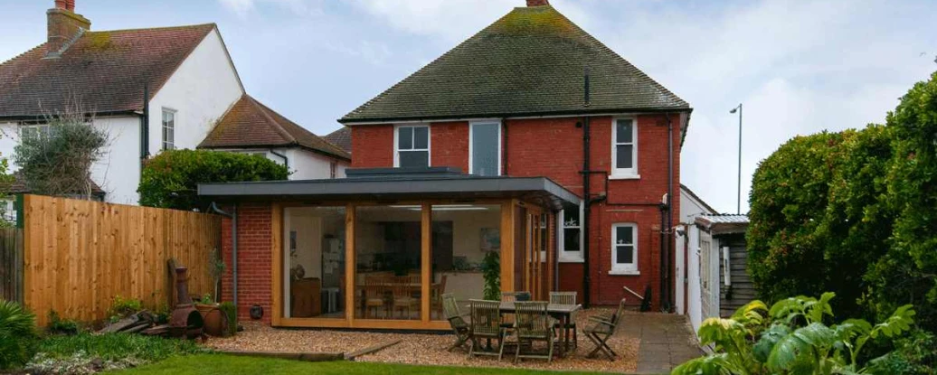 A red brick house with a modern glass extension in the backyard, surrounded by greenery and a wooden fence. The lawn is neatly manicured, and there is outdoor furniture on a paved area.