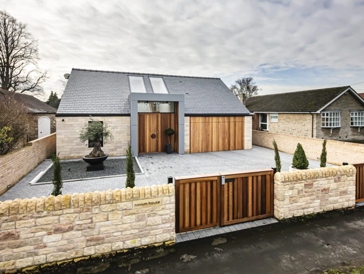A modern house featuring a stone façade and wooden accents. The entrance has a stylish door with a circular window above it. A landscaped front yard includes neatly trimmed shrubs and a decorative water feature. The driveway is paved, surrounded by a stone wall and wooden gates.
