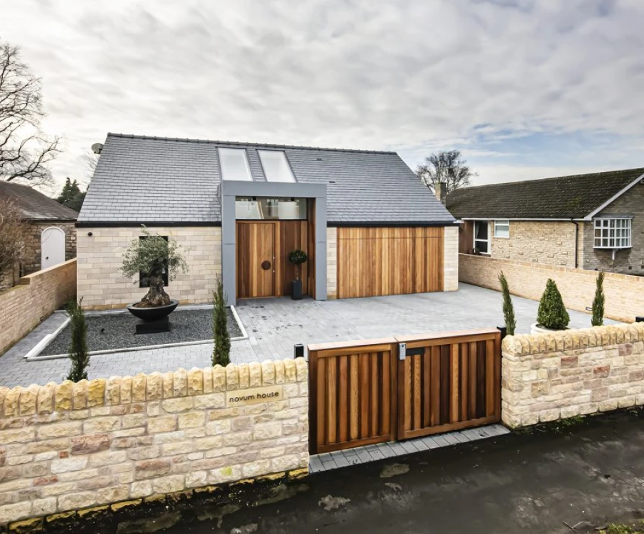 A modern house featuring a stone façade and wooden accents. The entrance has a stylish door with a circular window above it. A landscaped front yard includes neatly trimmed shrubs and a decorative water feature. The driveway is paved, surrounded by a stone wall and wooden gates.