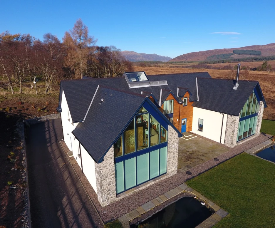 A modern house with a mix of stone and wood finishes, featuring large glass windows and a unique geometric roof design. The house is surrounded by a manicured lawn and a small pond, with rolling hills and trees in the background under a clear blue sky.