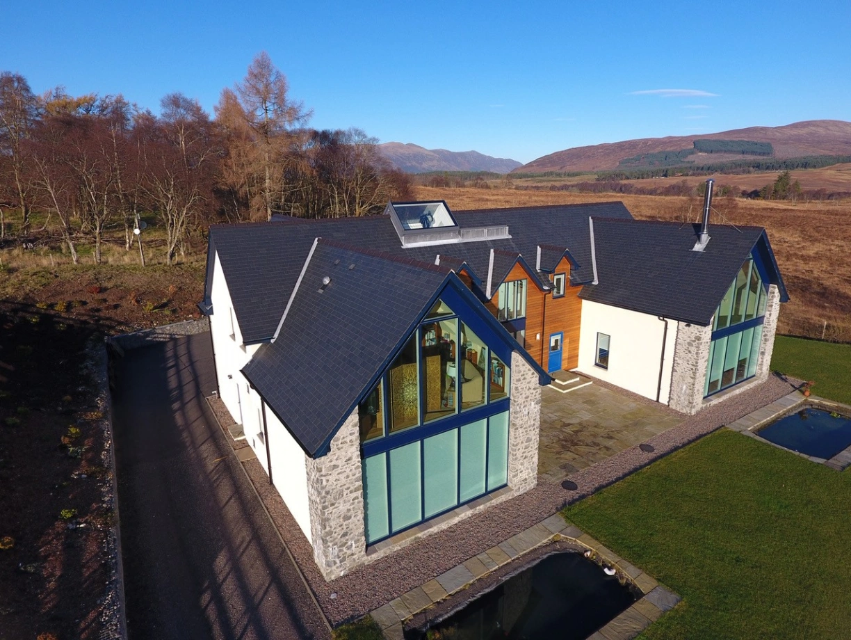 Casa moderna con un diseño arquitectónico distintivo, ubicada en un paisaje natural. Tiene grandes ventanales y un techo a múltiples aguas. Alrededor hay un jardín con césped y un pequeño estanque, con montañas al fondo.