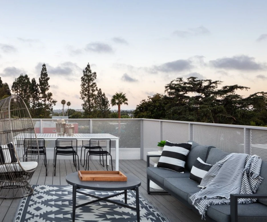A modern rooftop terrace featuring a cozy seating area with gray sofas and a black and white patterned throw blanket. A dining table with black chairs is set against a backdrop of lush trees and a colorful sunset sky.
