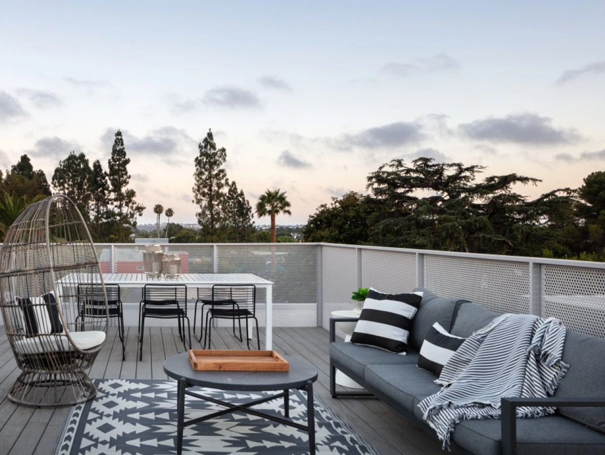 A modern rooftop terrace featuring a cozy seating area with gray sofas and a black and white patterned throw blanket. A dining table with black chairs is set against a backdrop of lush trees and a colorful sunset sky.
