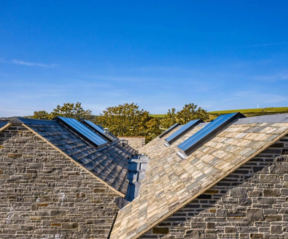 Aerial view of two sloped roofs made of slate tiles, featuring skylights. The backdrop shows a clear blue sky and green hills in the distance.