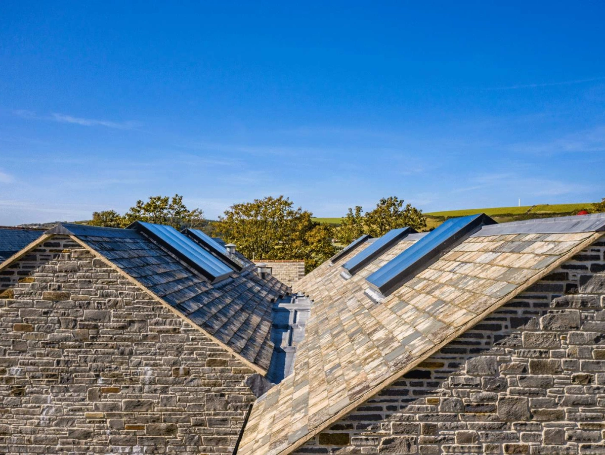 Two sloped rooftops covered in slate tiles, with skylights set into the slopes. Lush green hills are visible in the background under a clear blue sky.