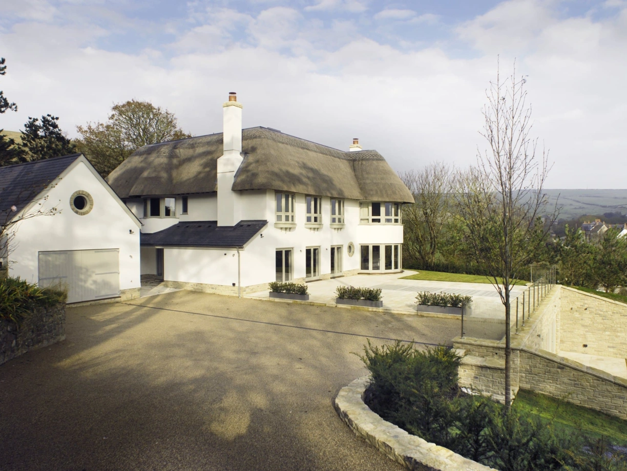 A modern white house with a thatched roof sits on a spacious driveway surrounded by trees. The landscape features a low stone wall and well-maintained gardens. Cloudy skies are visible in the background.