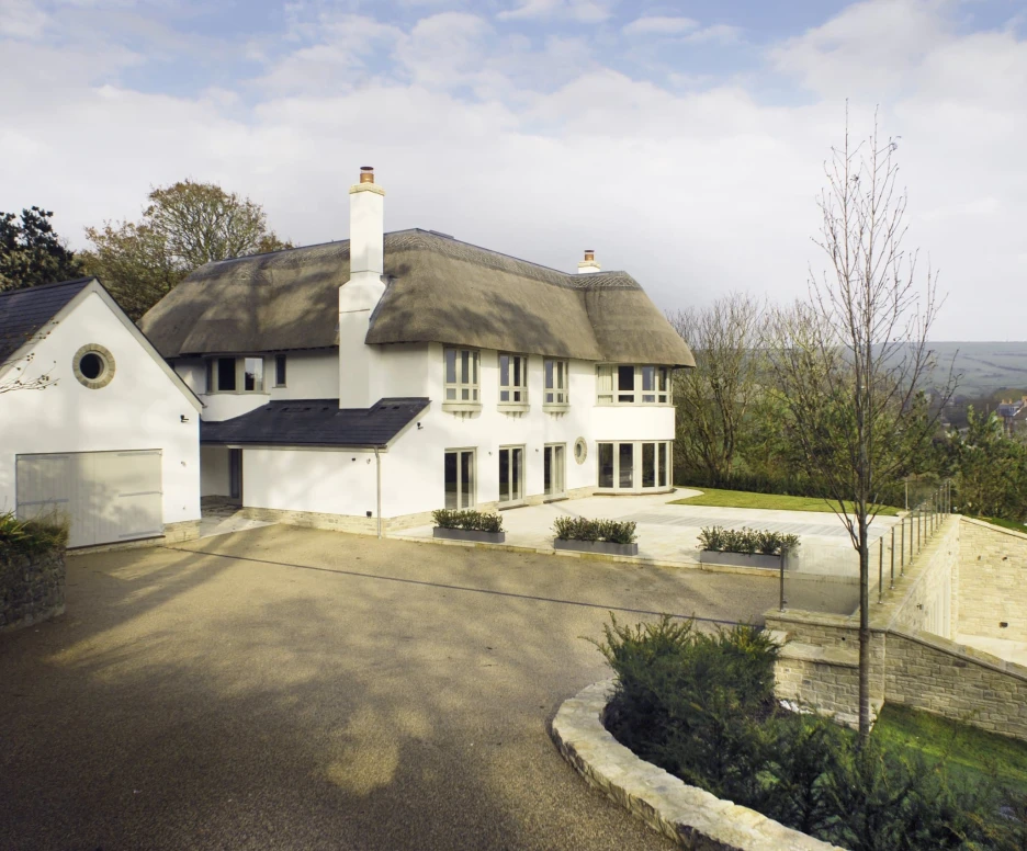 A modern white house with a thatched roof sits on a spacious driveway surrounded by trees. The landscape features a low stone wall and well-maintained gardens. Cloudy skies are visible in the background.