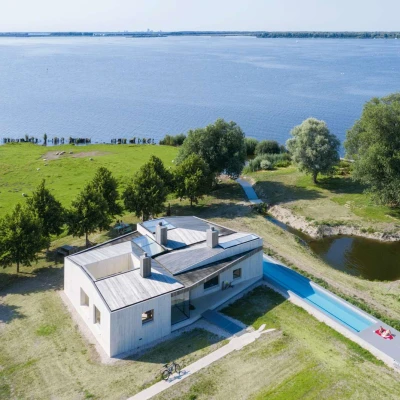 A modern house with a flat roof sits near a large body of water, surrounded by green grass and trees. A long, narrow swimming pool extends beside the house, leading to a serene landscape with calm water and distant wind turbines.