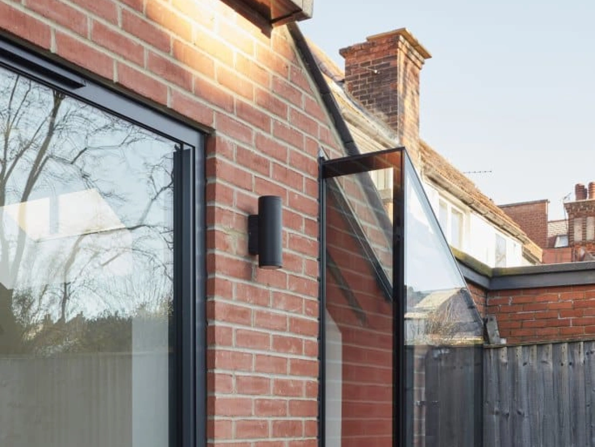 A modern brick exterior features large glass doors leading to a small balcony with a cozy seating area. The balcony, attached to a gray wall, has a sleek design and overlooks a fenced outdoor space.