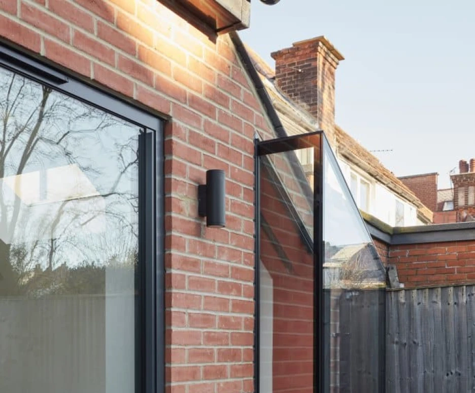 A modern brick exterior features large glass doors leading to a small balcony with a cozy seating area. The balcony, attached to a gray wall, has a sleek design and overlooks a fenced outdoor space.
