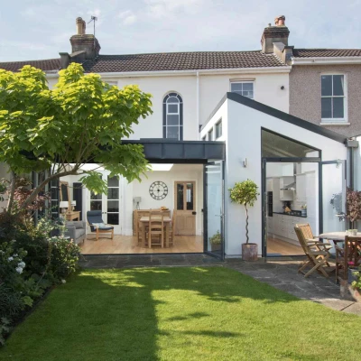 A modern home with a spacious garden features a glass extension leading to an open-plan interior. Lush green grass and colorful flowers surround a patio with wooden furniture. A large tree provides shade in the background.