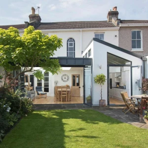 A modern home with a spacious garden features a glass extension leading to an open-plan interior. Lush green grass and colorful flowers surround a patio with wooden furniture. A large tree provides shade in the background.