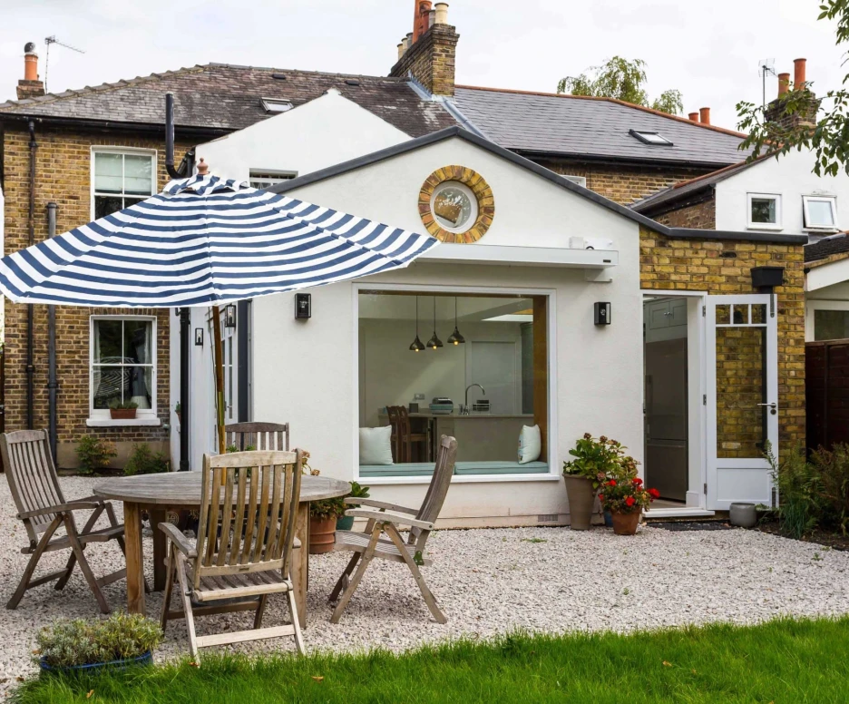 A cozy outdoor patio with wooden furniture, featuring a round dining table and striped umbrella. The background shows a charming white-walled house with a large window and a decorative clock. Lush green grass and plants enhance the inviting atmosphere.