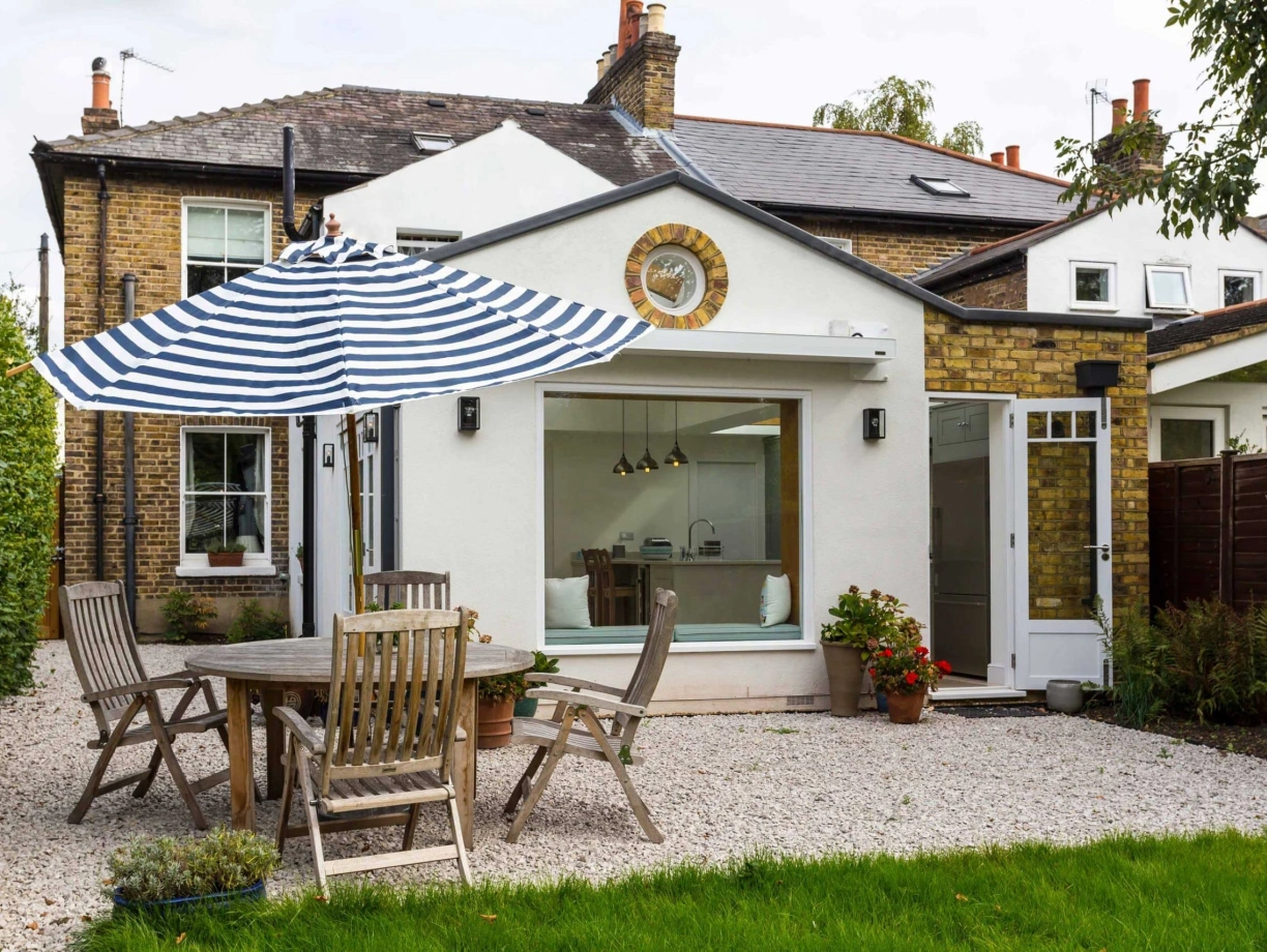 A cozy outdoor patio with wooden furniture, featuring a round dining table and striped umbrella. The background shows a charming white-walled house with a large window and a decorative clock. Lush green grass and plants enhance the inviting atmosphere.