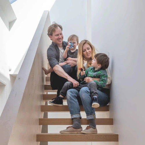 Eine Familie mit zwei kleinen Kindern sitzt auf einer Treppe in einem hellen Raum. Der Vater hält ein Kind, während die Mutter ein anderes Kind in den Armen hat. Alle lächeln und wirken entspannt. Die Treppe hat eine moderne Holzoberfläche und die Wände sind hell gestrichen.