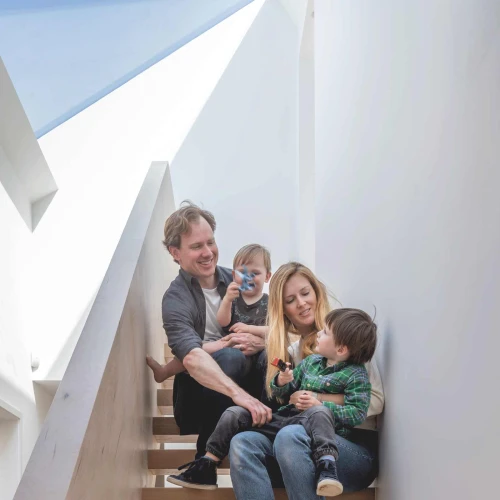 A family of four sits on a wooden staircase. The father holds a young child, while the mother sits with another child on her lap. All are smiling and engaged, with natural light coming from above.