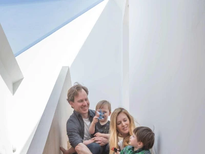 A family sits together on a staircase. The mother and father hold their children, who are smiling and playing. The setting features modern architecture with a bright, airy atmosphere.