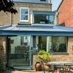 A modern single-story extension of a brick house features a glass wall and a blue metal roof. An outdoor table with chairs sits in front of the extension, surrounded by greenery.