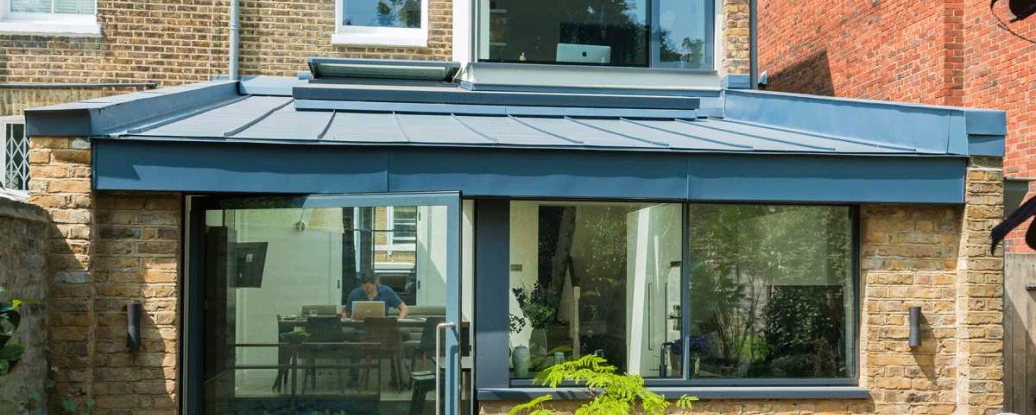 Modernized home extension with a blue metal roof and large glass doors. The structure features a blend of brick and stone, showcasing a spacious outdoor patio with a round table and plants. Sunlight filters in through large windows.