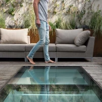 A person walks barefoot over a glass floor in a stylish outdoor living space, surrounded by modern sofas and greenery.