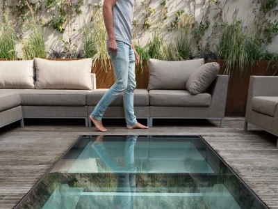 Ein Mann geht barfuß auf einem Glastreppenabsatz, umgeben von einer modernen Terrasse mit gemütlichen Sofas und grünen Pflanzen im Hintergrund.