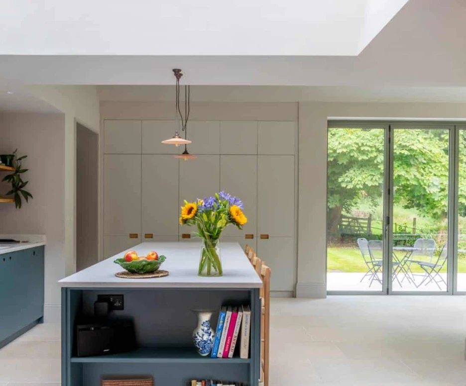 A modern kitchen featuring a central island with a blue base, a vase of sunflowers, and fresh fruits. The space is bright and airy, with large windows offering a view of a green outdoor area and a small table set outside. Stylish cabinetry and a minimalist design enhance the contemporary feel.