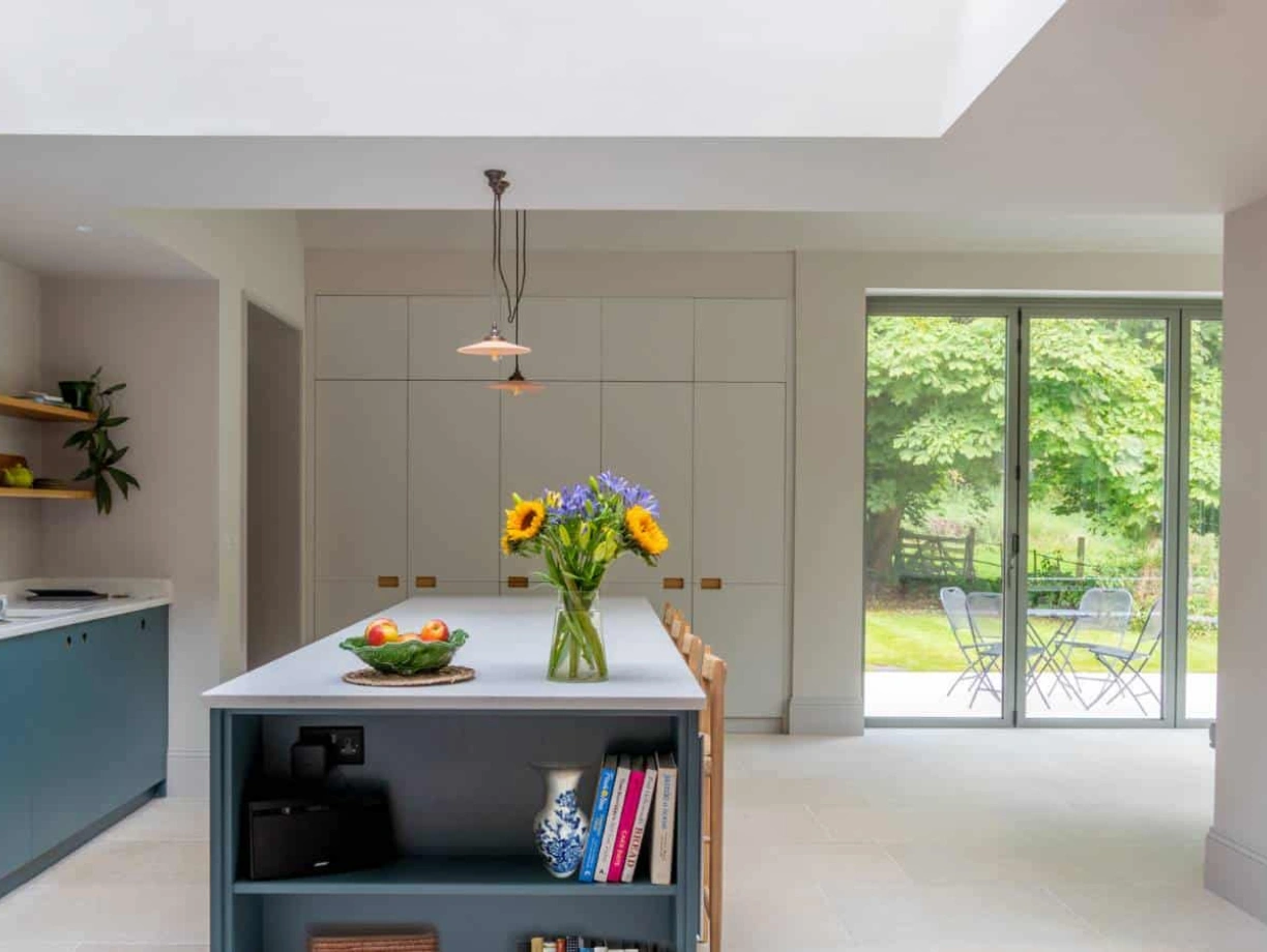 A modern kitchen featuring a central island with a blue base, a vase of sunflowers, and fresh fruits. The space is bright and airy, with large windows offering a view of a green outdoor area and a small table set outside. Stylish cabinetry and a minimalist design enhance the contemporary feel.