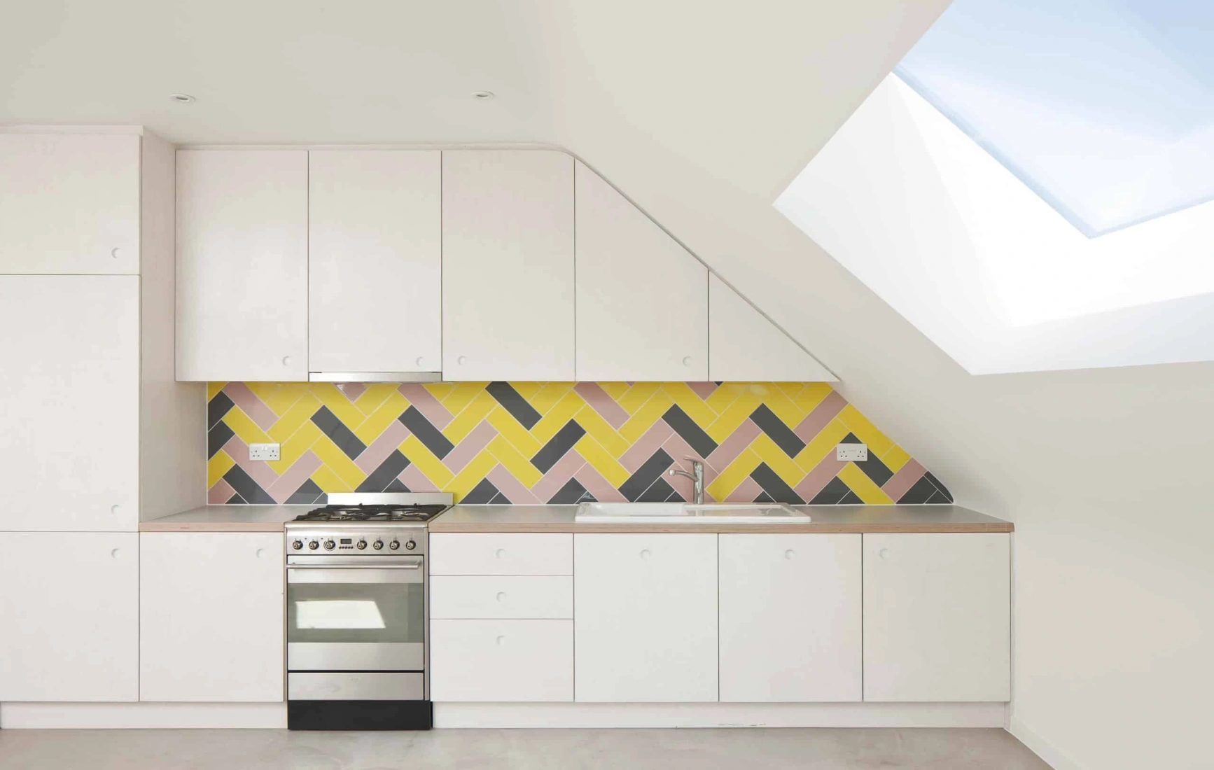 Modern kitchen featuring sleek white cabinetry, a stainless steel stove, and a colorful herringbone-patterned backsplash in shades of yellow, pink, and gray. A skylight adds natural light to the space.