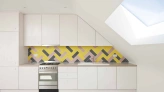 Cocina moderna con armarios blancos, una pared de azulejos en tonos amarillo, rosa y negro, y una ventana en el techo que permite la entrada de luz natural.